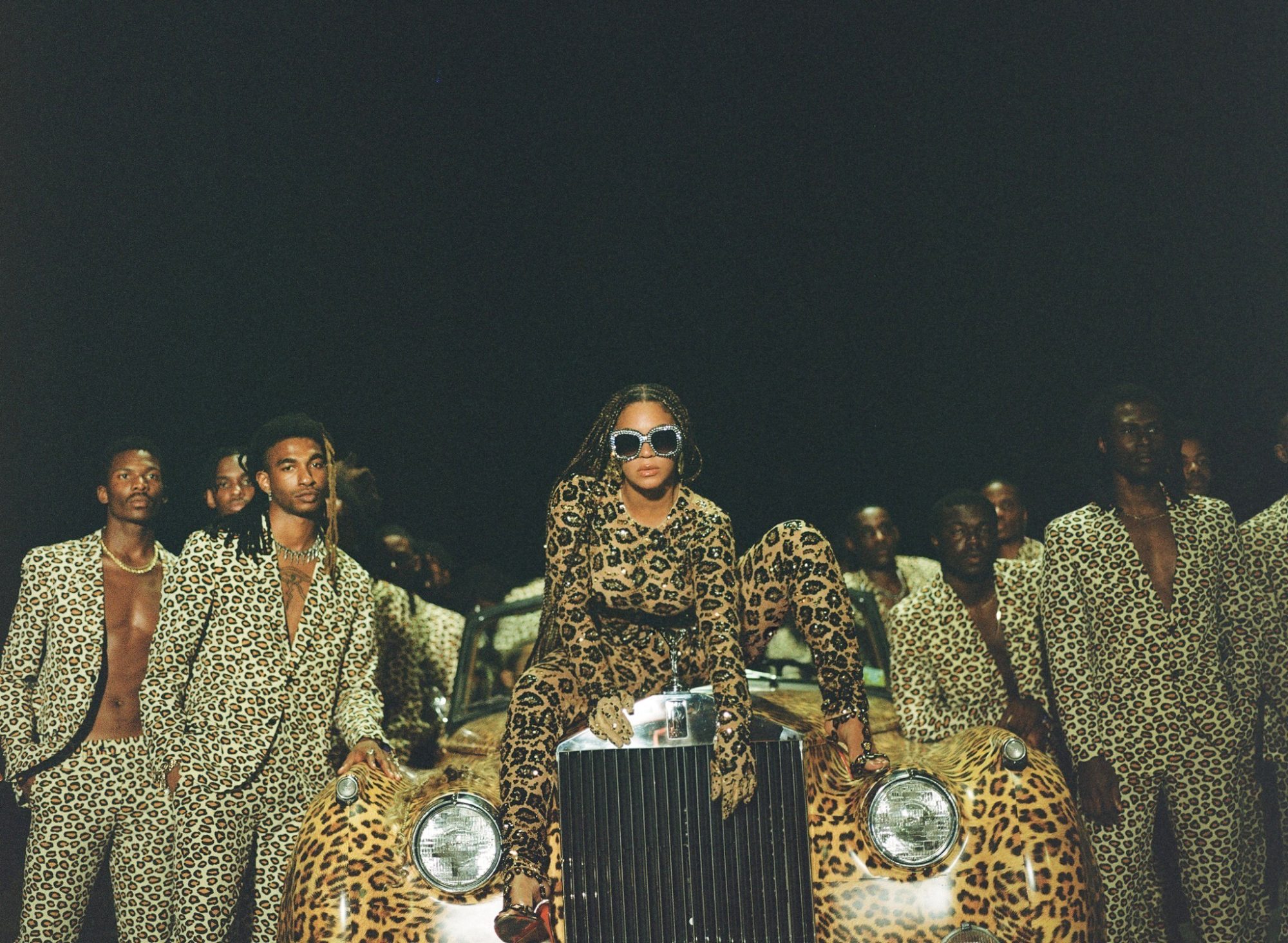 [Beyoncé, in a leopard print bodysuit, sits on a leopard print car surrounded by Black men in leopard print suits.]