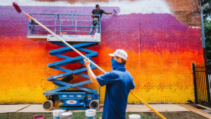 One man paints a wall standing on a crane, another man holds a paint roller